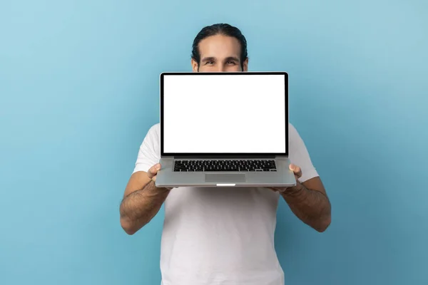 一个留着胡子的男人的画像 穿着白色的T恤 在笔记本电脑后面用白色的空显示屏遮住半张脸 用和善的眼睛看着相机 室内拍摄被蓝色背景隔离 — 图库照片
