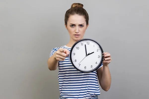 Portrait of bossy serious woman wearing striped T-shirt pointing finger at you holding in hand big wall clock, motivation. Indoor studio shot isolated on gray background.