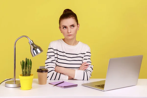 在办公室的笔记本电脑上工作时 穿着条纹衬衫的忧心忡忡的女人 带着沉思而困惑的表情坐在那里 对项目进行思考 在黄色背景下隔离的室内工作室拍摄 — 图库照片