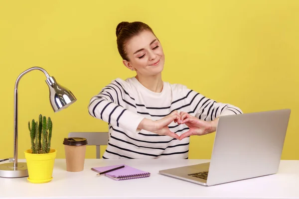 ノートパソコンで職場に座っているノートパソコンの画面に手で心のジェスチャーを示す積極的な女性は ロマンチックな感情を表現笑顔 黄色の背景に隔離された屋内スタジオショット — ストック写真