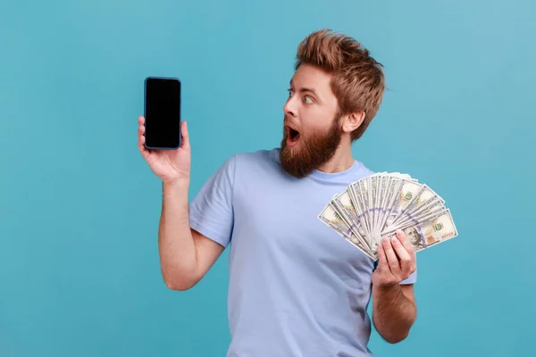 驚くべき驚きハンサムなひげを生やした男の肖像画は ドル紙幣を保持し 空の画面で携帯電話を見て オンライン賭け 青い背景に隔離された屋内スタジオショット — ストック写真