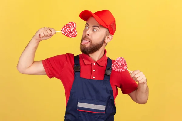形象滑稽的饥饿工人头戴红帽子 身穿蓝色制服 手里拿着美味的糖糖 舔着心形棒棒糖 伸出舌头 在黄色背景下隔离的室内拍摄 — 图库照片