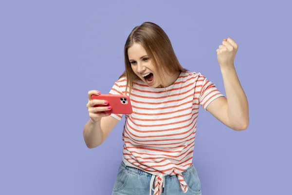 形象的兴奋微笑的女性游戏玩家穿着条纹T恤在智能手机上玩游戏 赢得水平 握紧拳头 庆祝她的胜利 室内拍摄被紫色背景隔离 — 图库照片