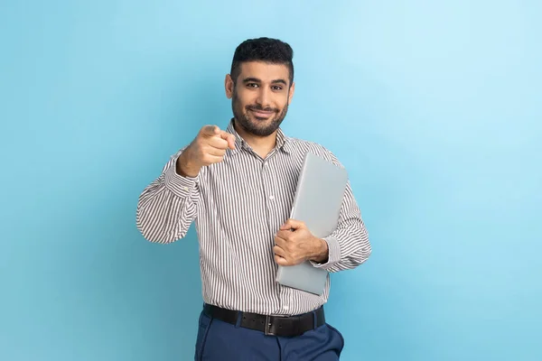一个快乐的商人站在那里 拿着封闭的笔记本电脑或文件夹 带着积极的表情 指着相机 穿着条纹衬衫 室内拍摄被蓝色背景隔离 — 图库照片