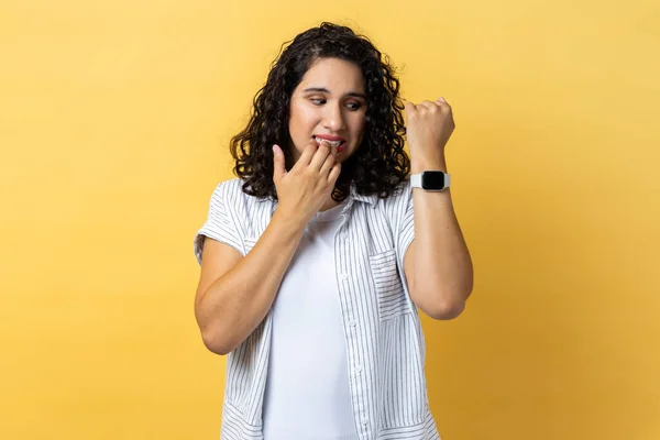 Portrait of displeased nervous woman with dark wavy hair biting finger nails and looking at her wrist clock with anxious, worried about deadline. Indoor studio shot isolated on yellow background.