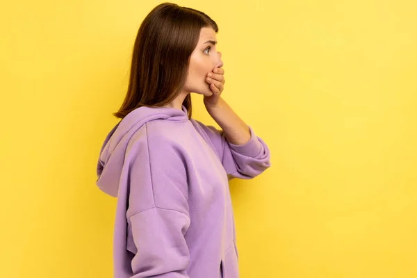 ショックを受けた暗い髪の女性のサイドビューの肖像画口の中に手を置き 先を見て 恐ろしい秘密を保ち 紫色のパーカーを着て 黄色の背景に隔離された屋内スタジオショット — ストック写真