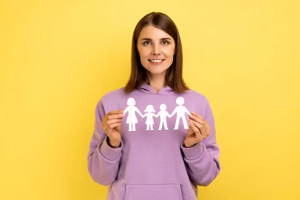 Kind smiling positive woman holding paper chain people which holding in hands, happy family, relationships, parenthood, wearing purple hoodie. Indoor studio shot isolated on yellow background.