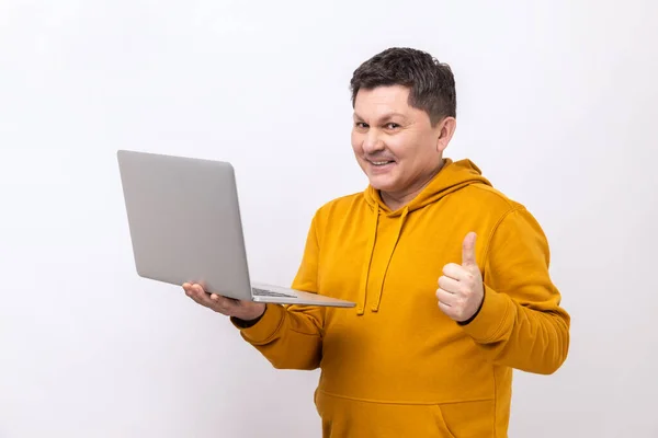 積極的な笑顔の男のフリーランサー親指を立って ノートパソコンを保持し テレワークに満足し 彼の仕事のように 都会的なスタイルのパーカーを着て表示されます 白い背景に隔離された屋内スタジオショット — ストック写真