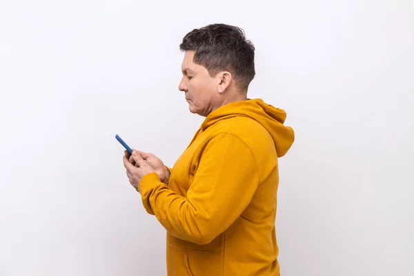 通过手机在社交媒体上发短信 使用移动网络服务 穿着都市风格的帽衫的中年男子的侧视图 在白色背景下被隔离的室内拍摄 — 图库照片