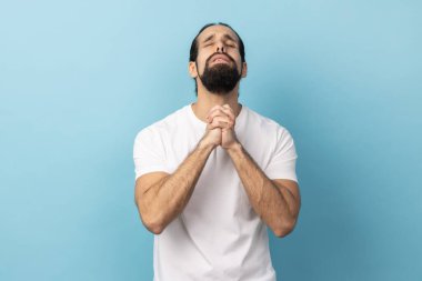 Man wearing white T-shirt looking up holding hands together, praying God, asking for health and support pleading for healing and forgiveness. Indoor studio shot isolated on blue background.