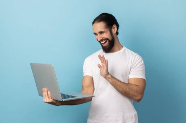 Portrait of smiling delighted man with beard wearing white T-shirt having video call, waving hand to camera, saying hello or good bye. Indoor studio shot isolated on blue background.