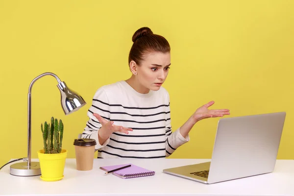 Young woman office worker raising hands in indignant gesture, asking why and looking angrily while having video call with business partner. Indoor studio studio shot isolated on yellow background.