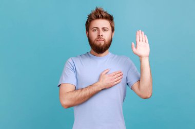 Portrait of honest sincere bearded man giving promise with hand on heart, pledging allegiance, giving vow with responsible serious face. Indoor studio shot isolated on blue background.