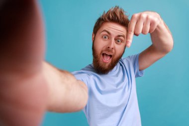 Portrait of bearded man taking selfie or making video call, looking at camera POV, point of view of photo, pointing finger down, subscribe. Indoor studio shot isolated on blue background.