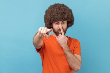 Portrait of man with Afro hairstyle wearing orange T-shirt touching nose, showing liar gesture, angry about falsehood, pointing to camera fake news. Indoor studio shot isolated on blue background.