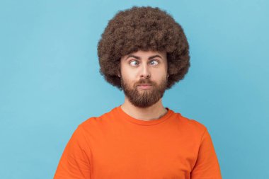 Portrait of crazy funny man with Afro hairstyle wearing orange T-shirt standing with crossed eyes and looking with silly comedian face. Indoor studio shot isolated on blue background. clipart
