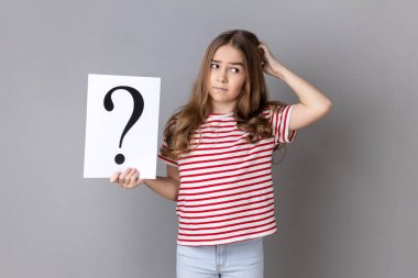 Portrait of little girl wearing striped T-shirt holding paper with question mark over, thoughtful, face thinking about question, very confused idea. Indoor studio shot isolated on gray background. clipart