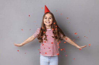 Portrait of charming extremely happy little girl wearing striped T-shirt standing with spread arms under falling confetti, celebrating birthday. Indoor studio shot isolated on gray background.