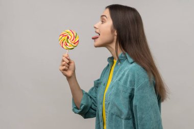 Side view of childish woman licking multicolor candy, wants to eat, looking at camera, showing tongue out, wearing casual style jacket. Indoor studio shot isolated on gray background.