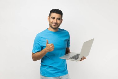Portrait of happy delighted attractive unshaven man wearing blue T- shirt standing holding laptop in hands, working online, showing thumb up. Indoor studio shot isolated on gray background.