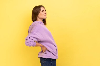 Profile portrait of unhealthy frustrated anxious woman holding hands on lower back, kidney injury, backache, wearing purple hoodie. Indoor studio shot isolated on yellow background.