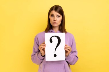 Portrait of serious concentrated woman looking at camera, holding paper with question mark, thinks about tasks, wearing purple hoodie. Indoor studio shot isolated on yellow background.