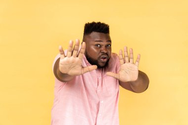No, it's scary. Portrait of frightened shocked man wearing pink shirt raising hands in fear, looking horrified and panicking, hiding from phobia. Indoor studio shot isolated on yellow background.