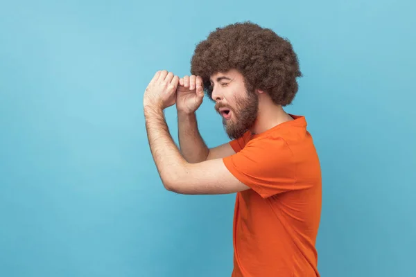 Side view of man with Afro hairstyle wearing orange T-shirt making glasses shape, looking through monocular gesture with amazed expression. Indoor studio shot isolated on blue background.