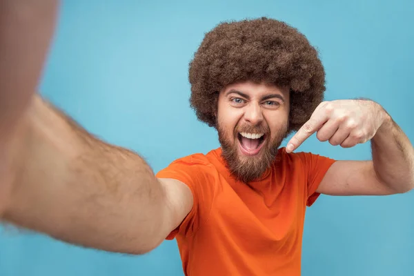Portrait of smiling satisfied man blogger with Afro hairstyle wearing orange T-shirt broadcasting livestream, pointing down, asking to subscribe, POV. Indoor studio shot isolated on blue background.