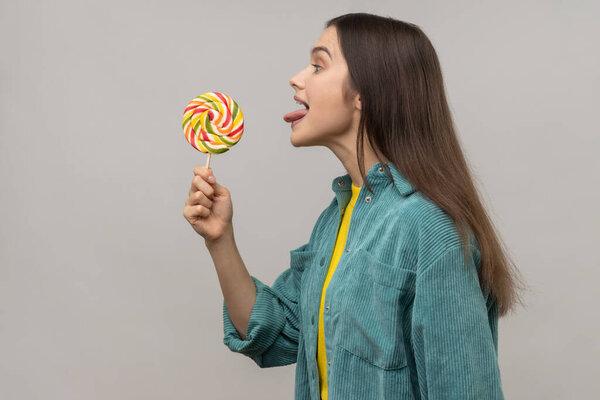 Side view of childish woman licking multicolor candy, wants to eat, looking at camera, showing tongue out, wearing casual style jacket. Indoor studio shot isolated on gray background.