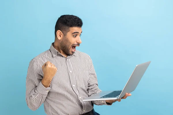Excited businessman with beard screaming yes raising hands up, solving hard task, getting access, successfully completing work, wearing striped shirt. Indoor studio shot isolated on blue background.