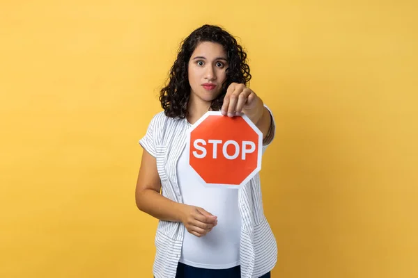 Portrait of serious woman with dark wavy hair holding red stop sign looking at camera with serious facial expression, prohibition. Indoor studio shot isolated on yellow background.