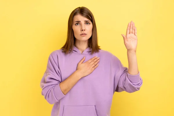 真剣に意識した女性は 胸に手を取り合い 紫色のパーカーを着て もう一つの誓う 信頼と正直 愛国心を高める 黄色の背景に隔離された屋内スタジオショット — ストック写真