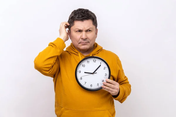 Portrait of middle aged man rubbing his head holding in hands big wallclock, has no time, worried about deadline, wearing urban style hoodie. Indoor studio shot isolated on white background.