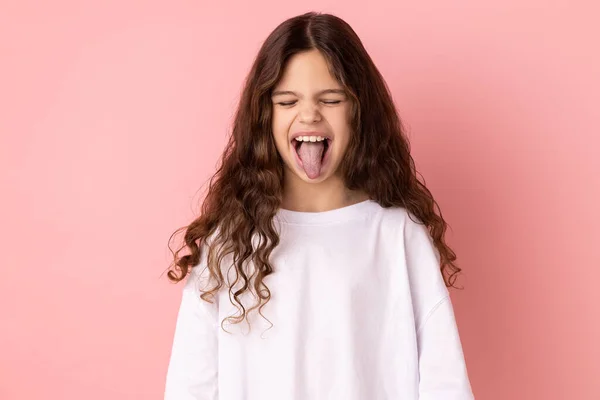 stock image Portrait of childish carefree little girl wearing white T-shirt showing out tongue and closing eyes with naughty disobedient grimace, making face. Indoor studio shot isolated on pink background.