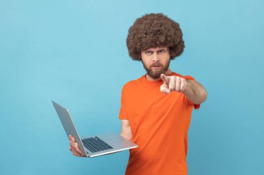 Strict bossy man with Afro hairstyle wearing orange T-shirt holding laptop in hands, working online, pointing finger to camera with serious expression. Indoor studio shot isolated on blue background.