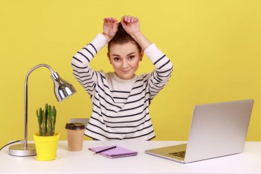Charming woman manager smiling and showing bunny ears on head, resting, having fun at workplace in home office, feeling carefree and happy. Indoor studio studio shot isolated on yellow background.