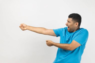 Side view portrait of strong attractive young adult unshaven man wearing blue T- shirt standing pulling, trying to pull something heavy. Indoor studio shot isolated on gray background.