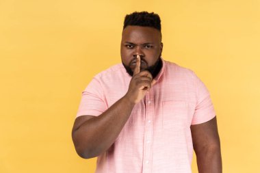 Shh, make silence please. Portrait of serious strict man wearing pink shirt standing showing quiet gesture, saying hush, keep secret. Indoor studio shot isolated on yellow background.