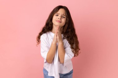 Please help me or forgive. Portrait of little girl wearing white T-shirt standing with palm hands and looking at camera pleased pleading. Indoor studio shot isolated on pink background.