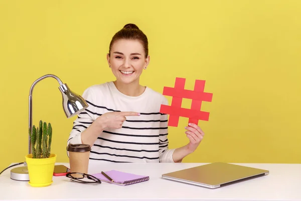 Young attractive woman sitting at workplace, pointing at big red hashtag symbol to laptop screen, sharing viral content, tagged message. Indoor studio studio shot isolated on yellow background.