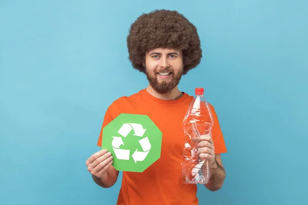 Portrait of smiling man with Afro hairstyle wearing orange T-shirt holding recycling green sign and plastic bottle, looking at camera. Indoor studio shot isolated on blue background.