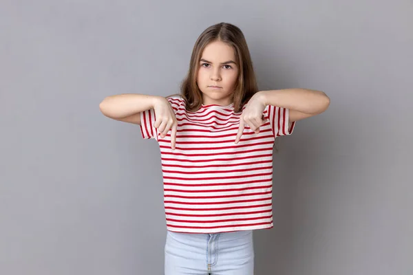 Here and right now. Portrait of serious bossy dark haired little girl wearing striped T-shirt standing and pointing index fingers down. Indoor studio shot isolated on gray background.