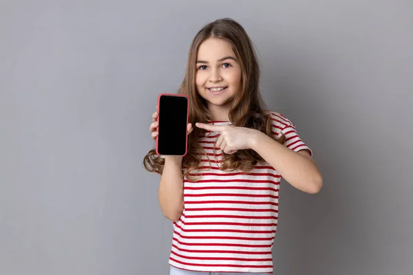 Portrait Delighted Little Girl Wearing Striped Shirt Pointing Finger Smartphone — Stockfoto