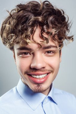 Portrait of attractive man with beard and mustache with wavy hairstyle, wearing blue shirt, looking at camera with toothy smile. Indoor studio shot isolated on gray background.