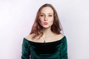 Portrait of beautiful gently young adult woman wearing green dress standing with pout lips, blowing air kissing, looking at camera. Indoor studio shot isolated on gray background.