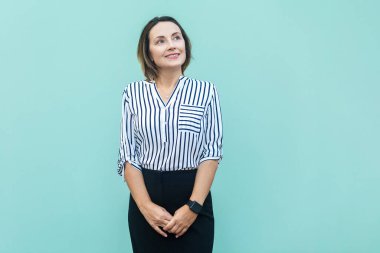 Portrait of positive smiling middle aged woman wearing striped shirt looking away, dreaming about future, having friendly pleased expression. Indoor studio shot isolated on light blue background.