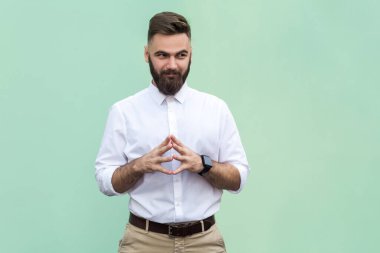 Portrait of attractive cunning bearded businessman wearing white shirt schemes something, keeps fingers together, considers over cunning plans. Indoor studio shot isolated on light green background.