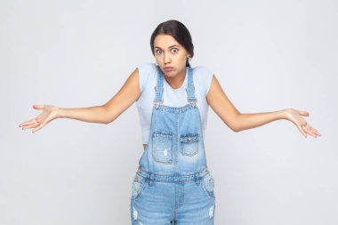 Maybe, don't know. Portrait of unsure woman wearing denim overalls raising hands with confused perplexed puzzled expression, not sure about decision. Indoor studio shot isolated on gray background.