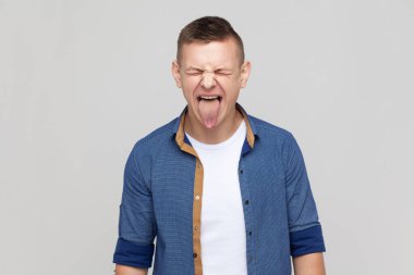 Childish disobedient behavior. Portrait of attractive teenager guy wearing blue shirt sticking tongue out, making naughty goofy grimace, teasing. Indoor studio shot isolated on gray background.